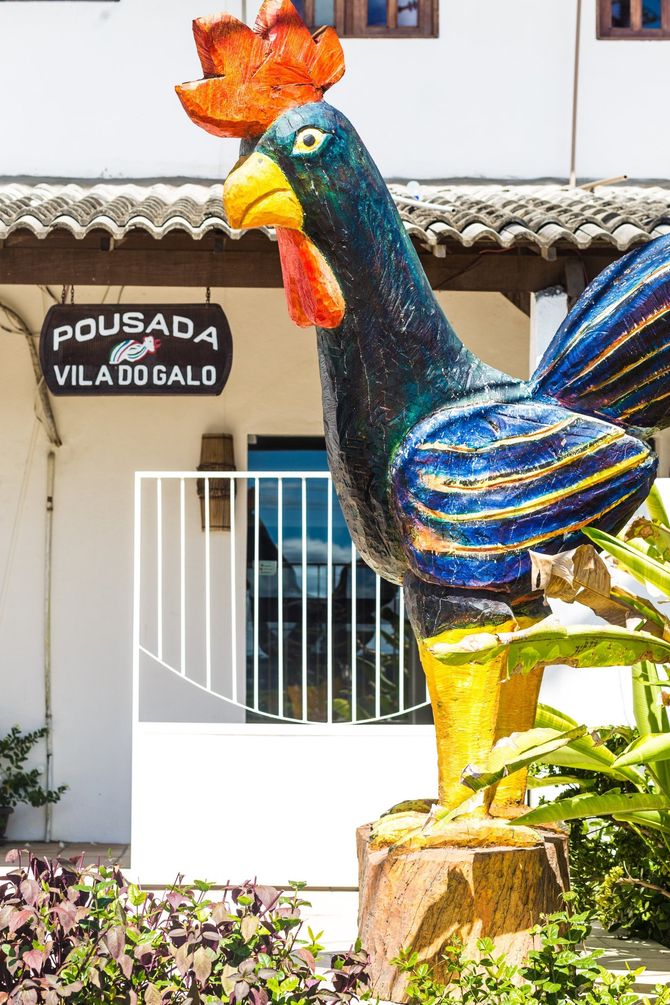 Estátua colorida de galo em frente à Pousada Vila do Salão, com o prédio branco e plantas do jardim ao fundo.