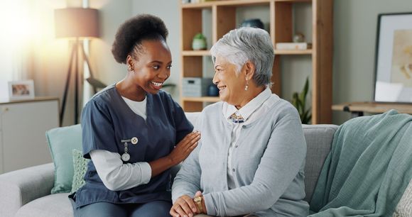 Caregiver with patient, smiling on a couch in a living room. Caregiver has arm around patient.