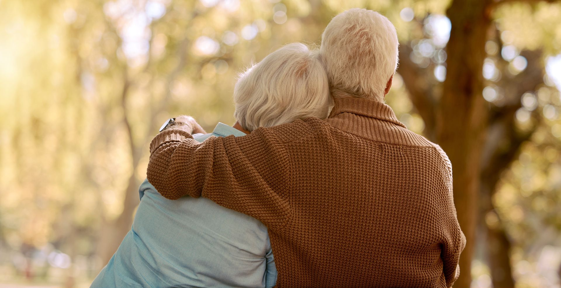 Two people in sweaters embracing outdoors in warm sunlight among trees.