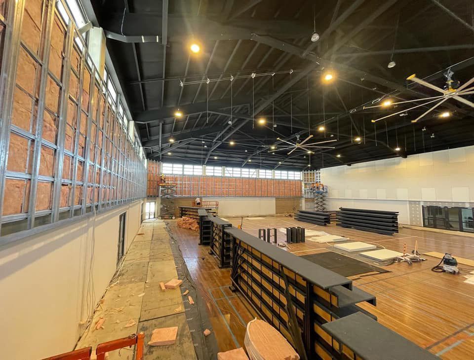 Interior of a large building under construction. Wall with metal framing, exposed insulation. Black ceiling, wood floor.