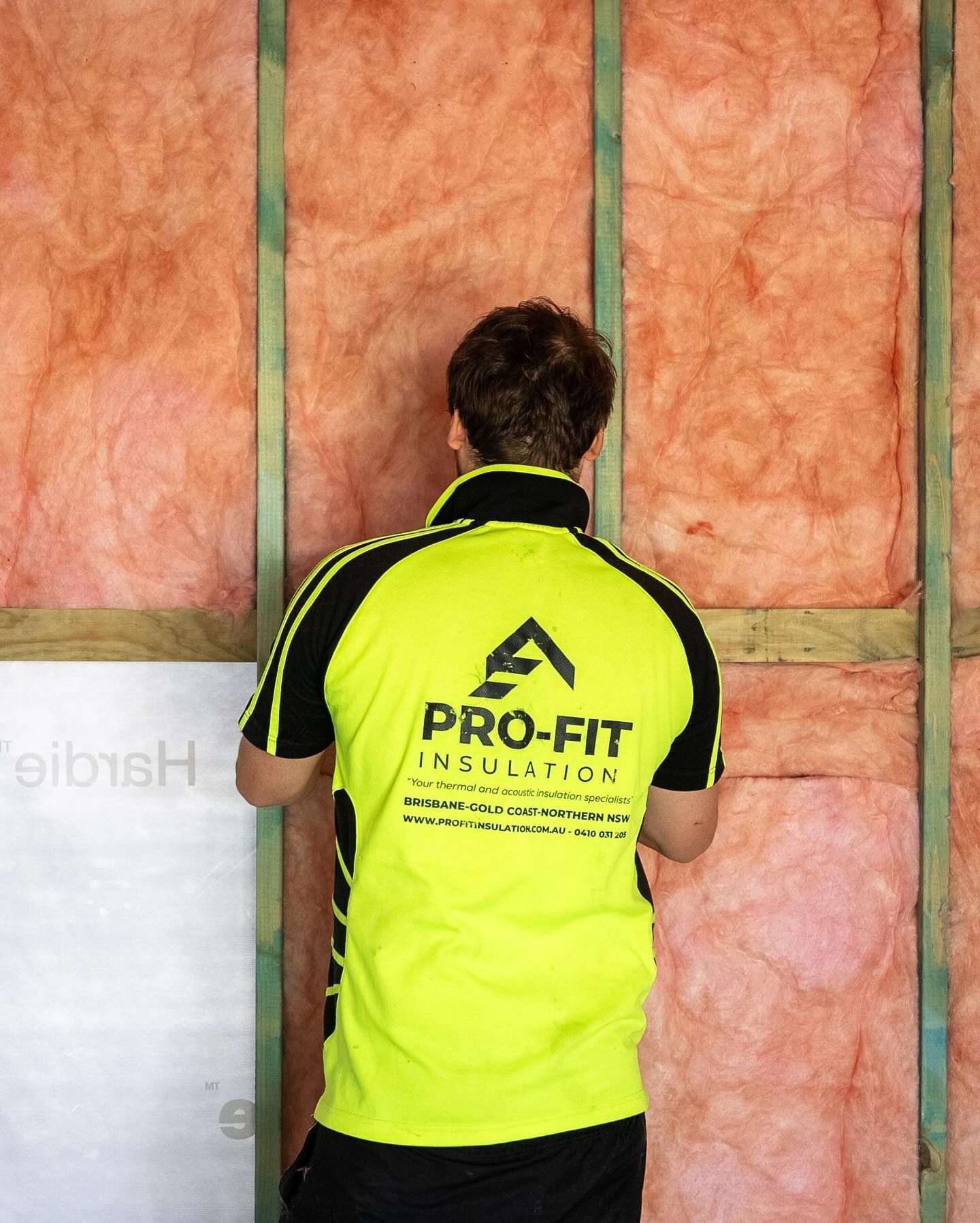 Man installing pink insulation, wearing a yellow Pro-Fit Insulation shirt, inside a wooden framed wall.