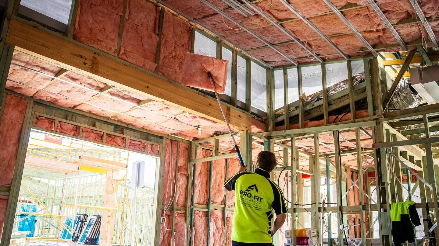 Construction worker installing insulation in a house frame, using a long handled tool.