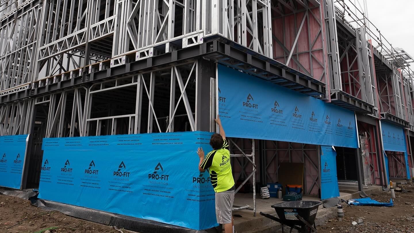 Construction worker installing blue weather-resistant barrier on a building's steel frame exterior.
