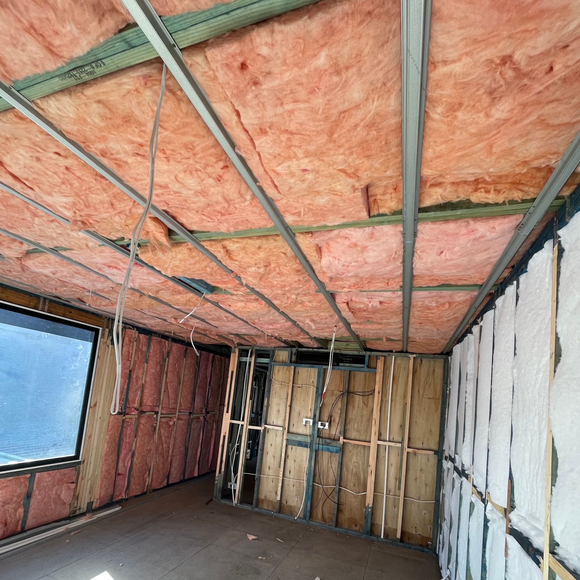 Interior of a room under construction with pink insulation in ceiling and walls, exposed framing and wiring.
