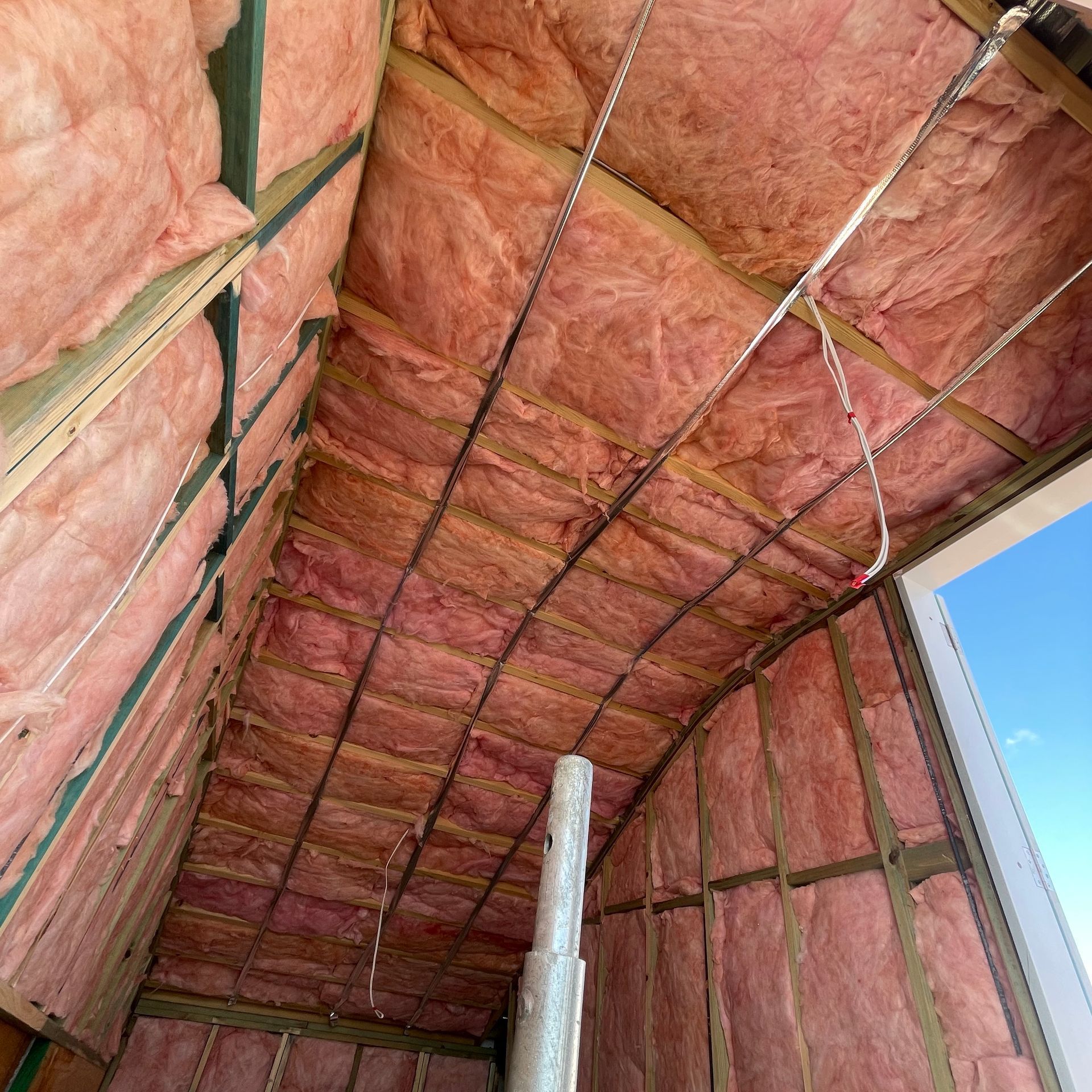 Interior view of a building under construction, featuring pink insulation between wooden beams; wires and a pipe are visible.
