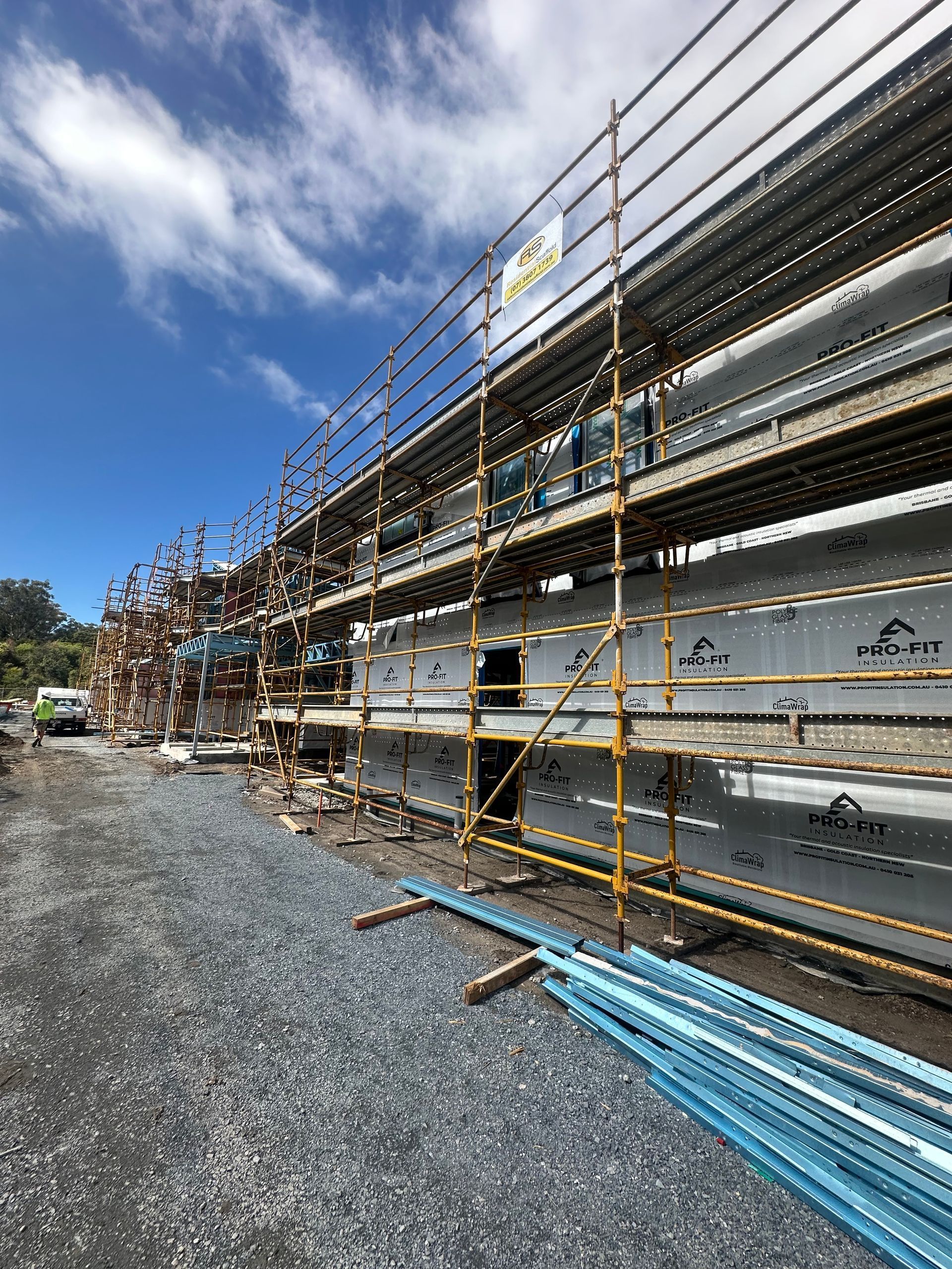 Scaffolding around a building under construction. Grey siding, blue sky, gravel path.