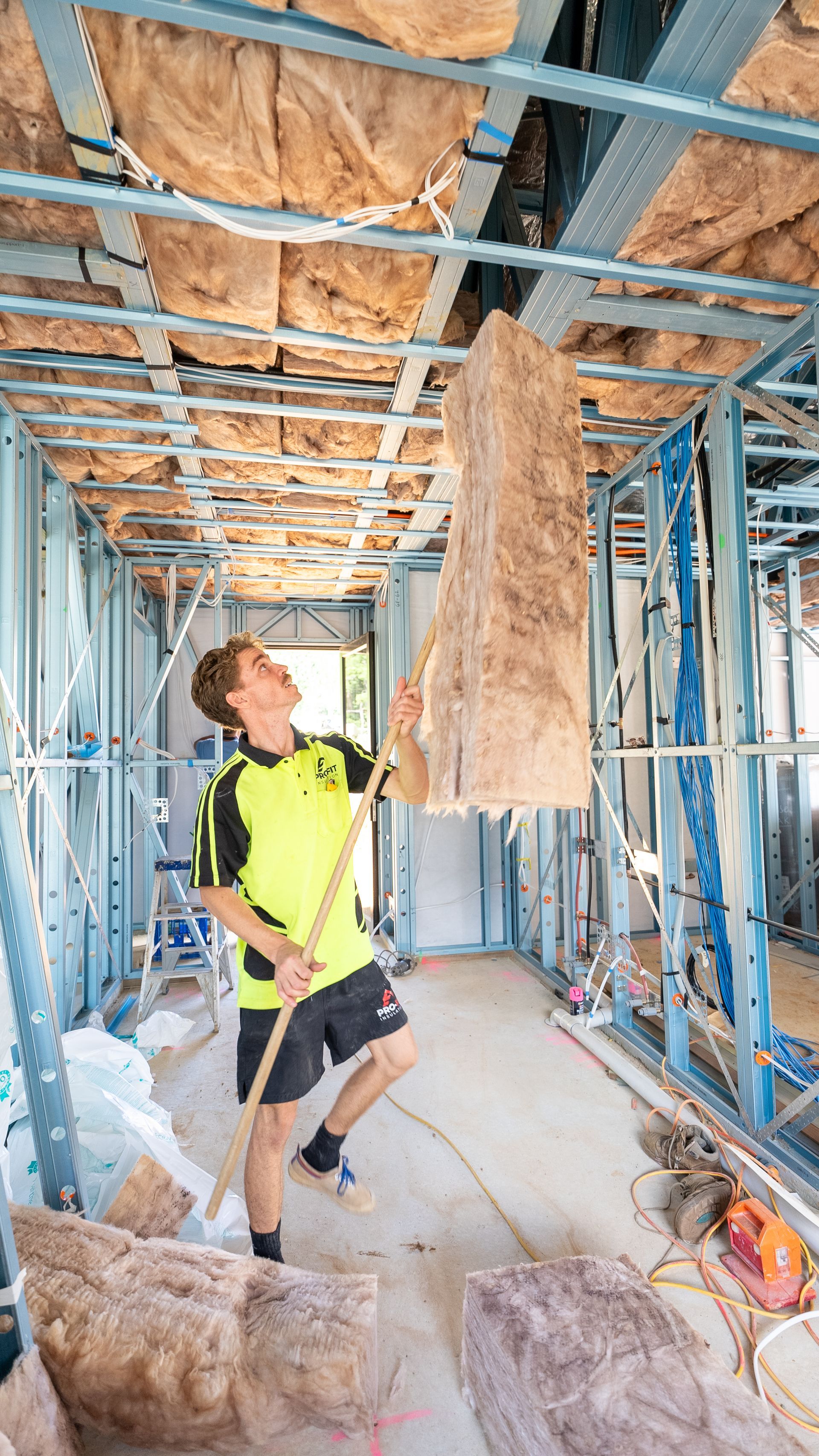 Construction worker installing insulation in a new building's ceiling, holding a large panel up with a pole.
