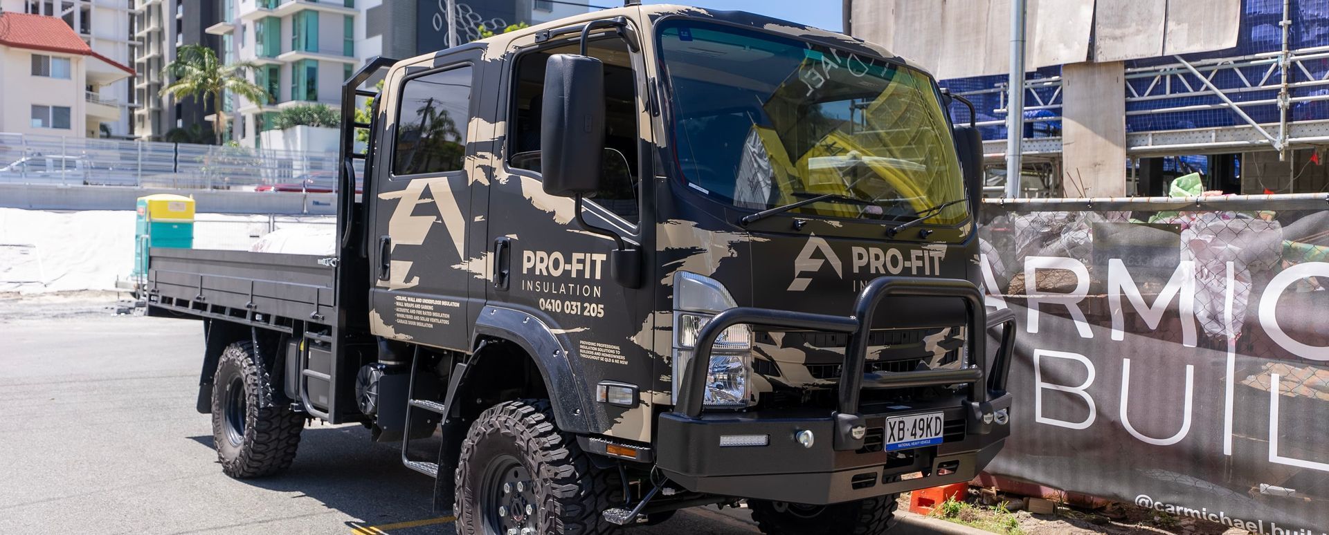 Camouflage-wrapped truck with flatbed, parked in front of a construction site with