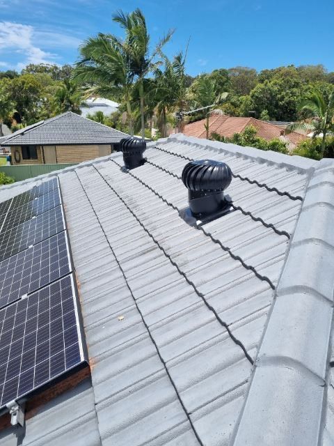 Rooftop with solar panels and two black turbine vents on gray tile roof. Green trees and blue sky.