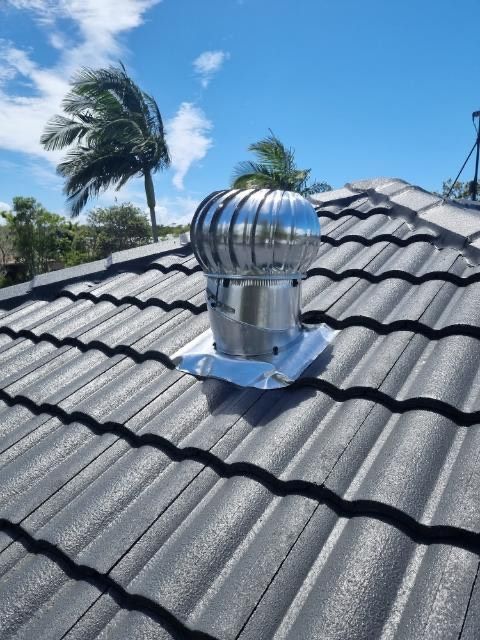 Silver roof vent on gray tile roof, palm tree in background, bright blue sky.