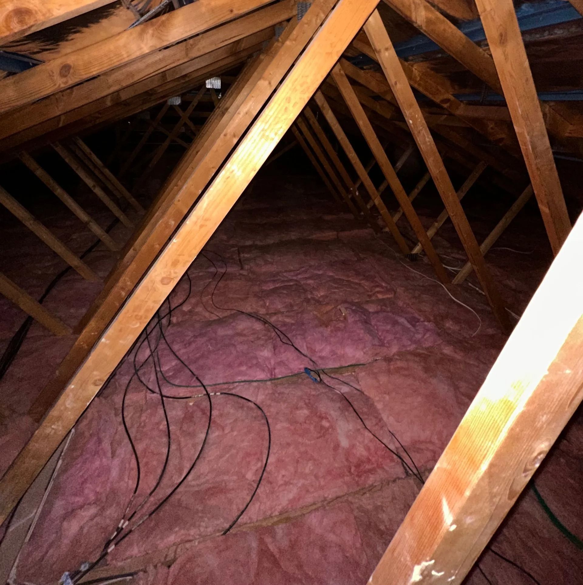 Attic interior with pink insulation and wooden beams. Black wires run along the insulation.