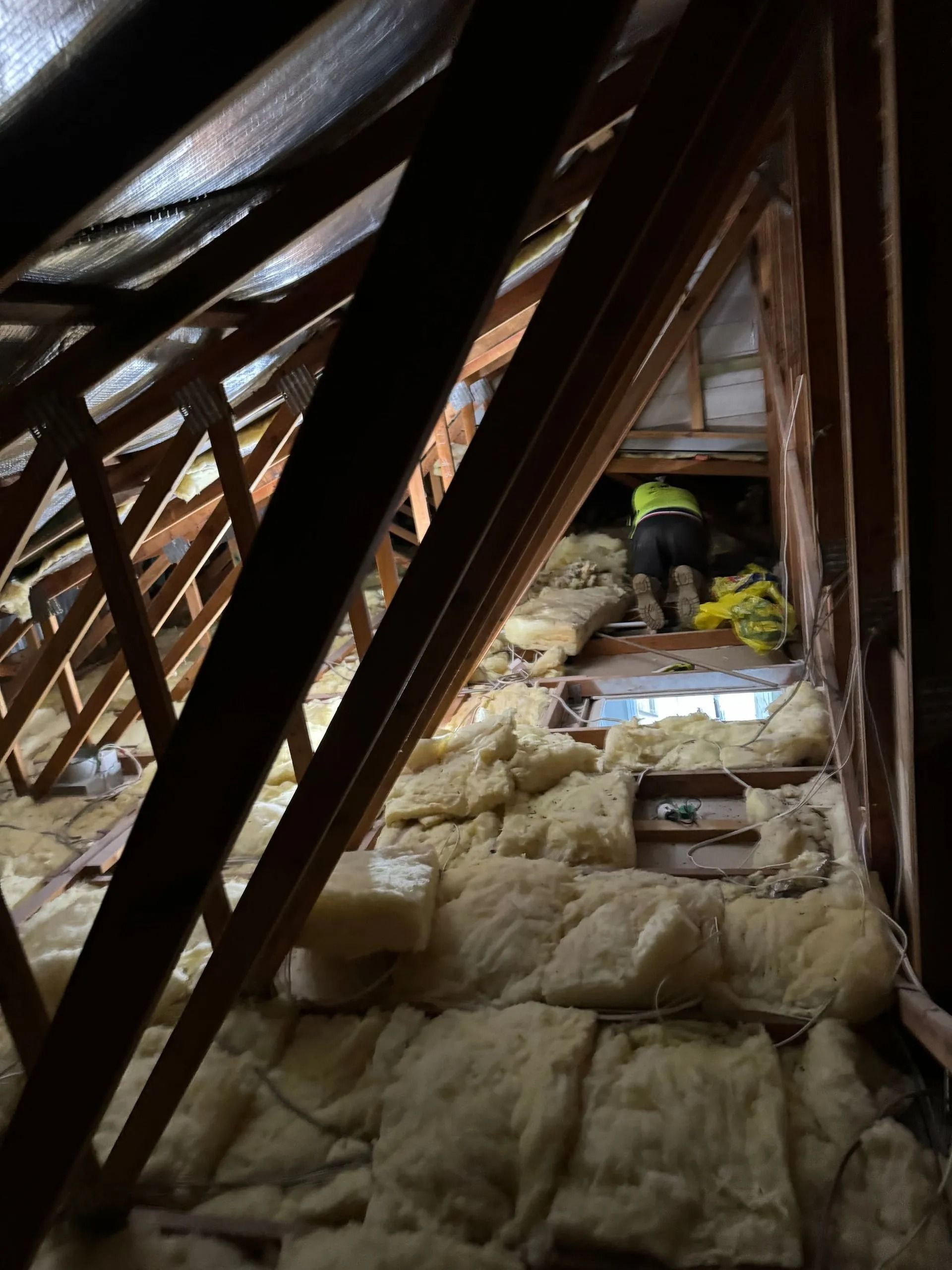 Attic interior with insulation and wooden beams. A person in a high-visibility vest is visible in the distance.