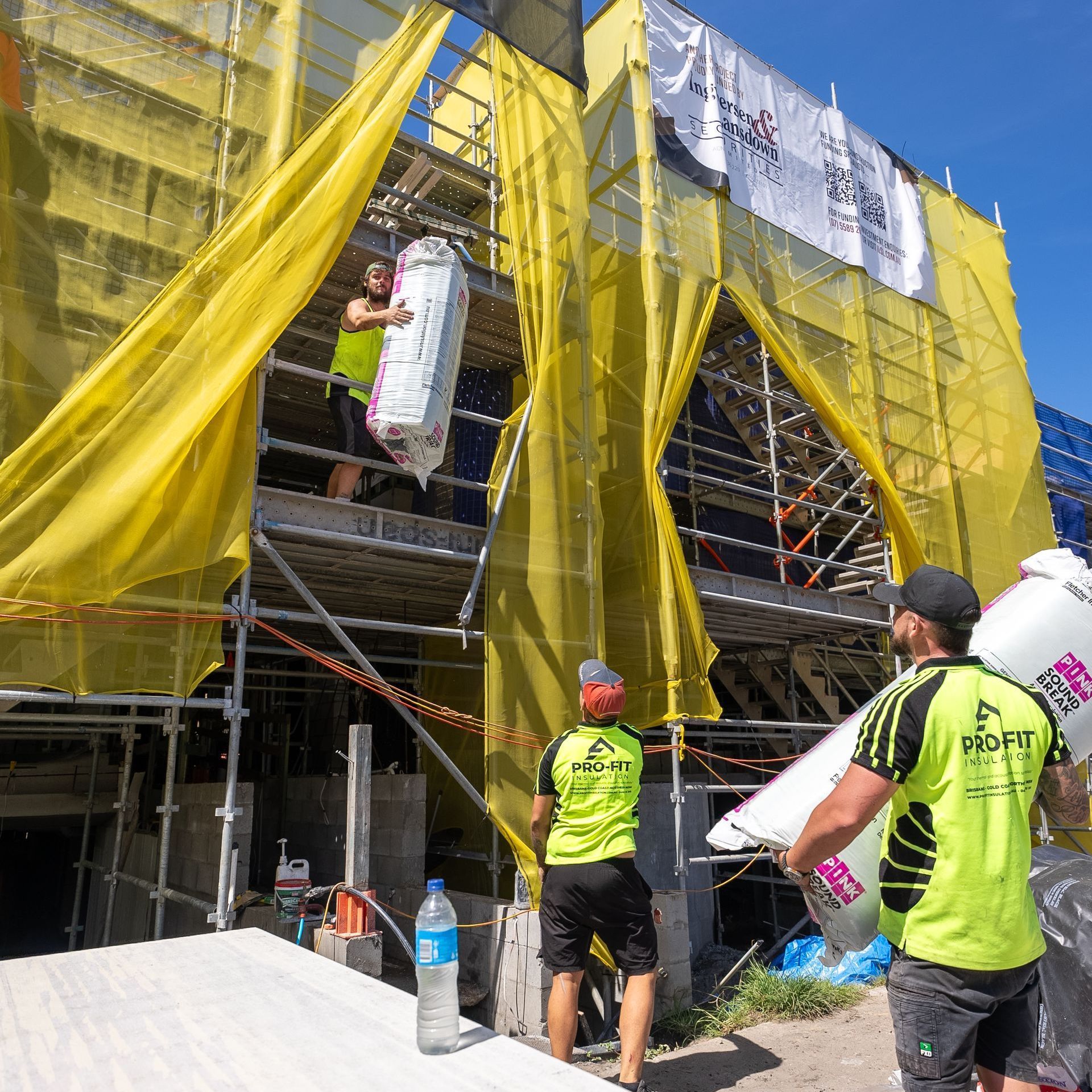 Construction workers on scaffolding, moving insulation at a building site; yellow safety netting.