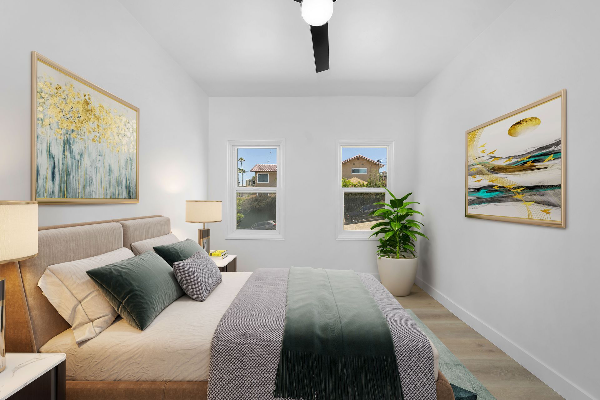 A bedroom with a large bed , two paintings on the wall and a ceiling fan.