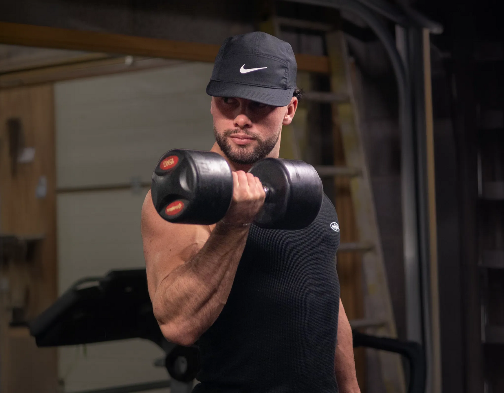 Man lifting a dumbbell in a dimly lit gym, wearing a Nike cap.