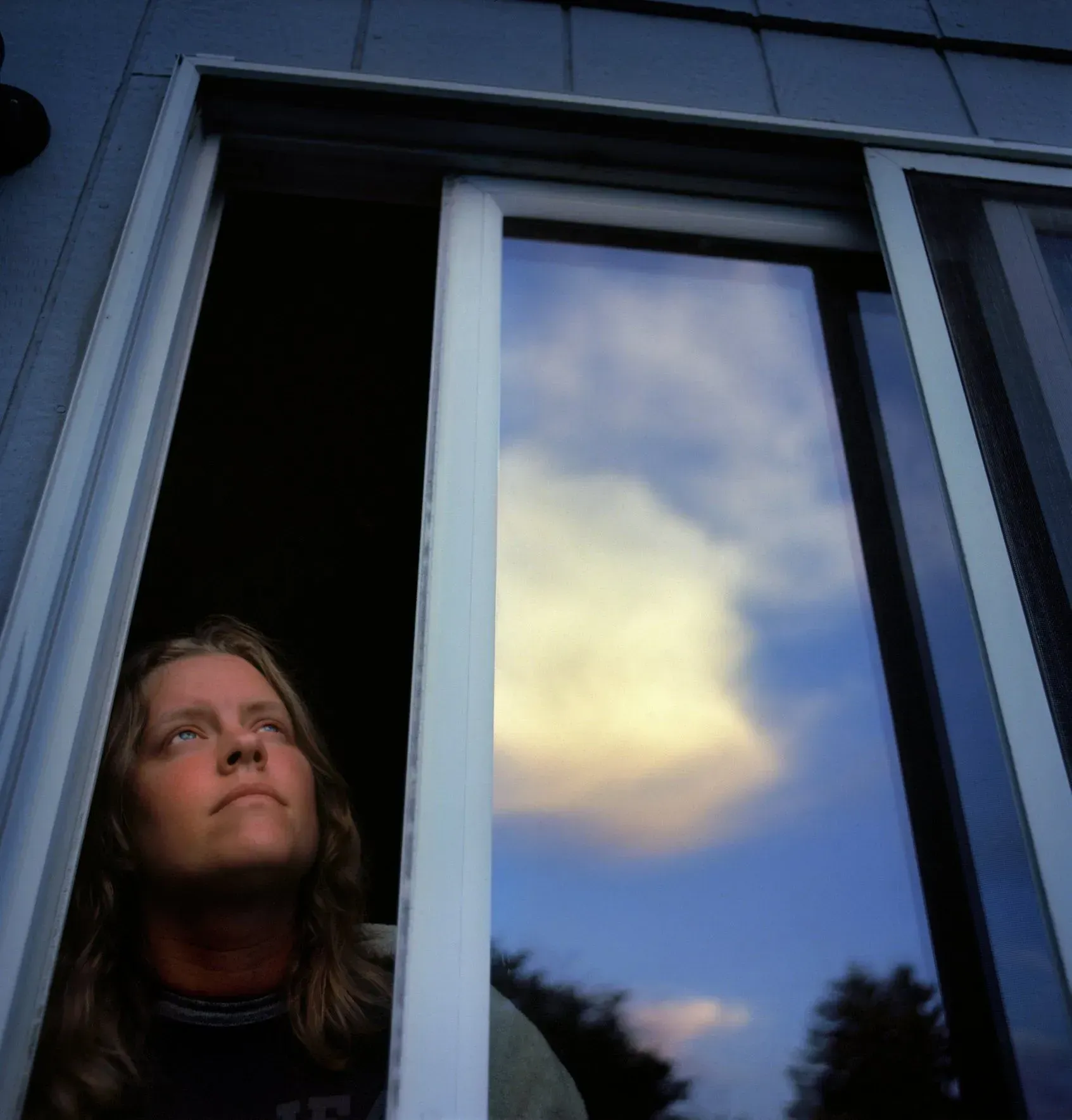 Person looking out from a dark doorway beside a reflective window at dusk