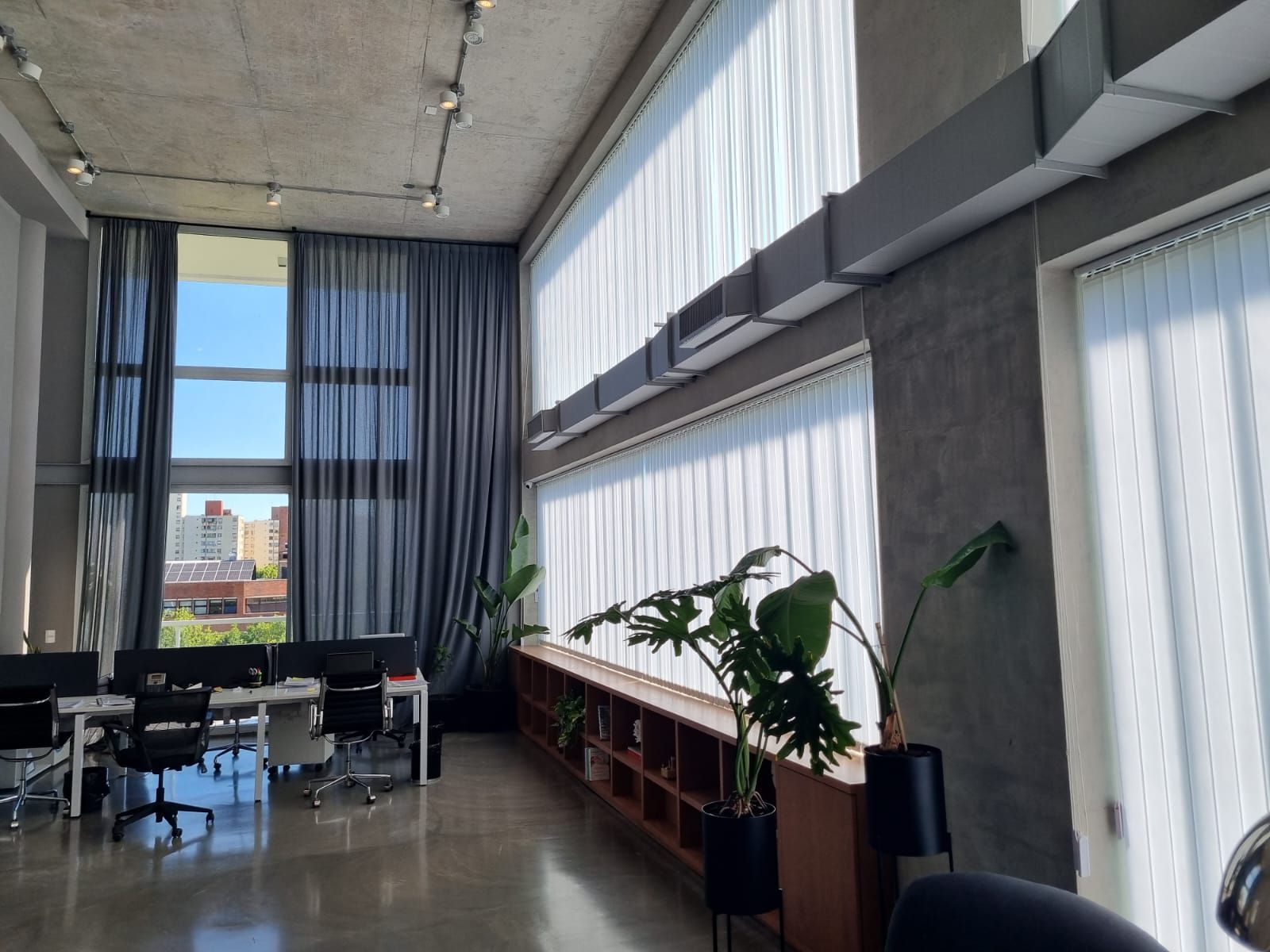 Oficina moderna y luminosa con grandes ventanales, cortinas, escritorios y plantas en macetas