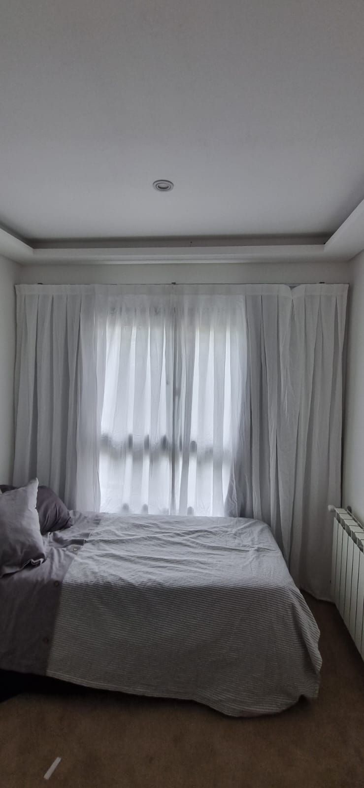 Un pequeño dormitorio con una cama, paredes grises, cortinas blancas translúcidas y luz natural suave.