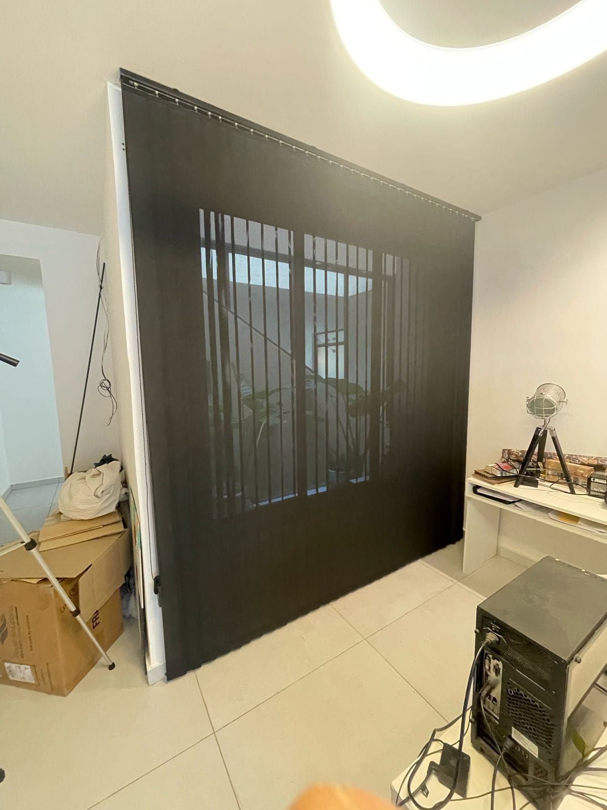Un gran panel corredizo negro o persiana apoyado contra una pared en una habitación luminosa con cajas y equipos cerca.