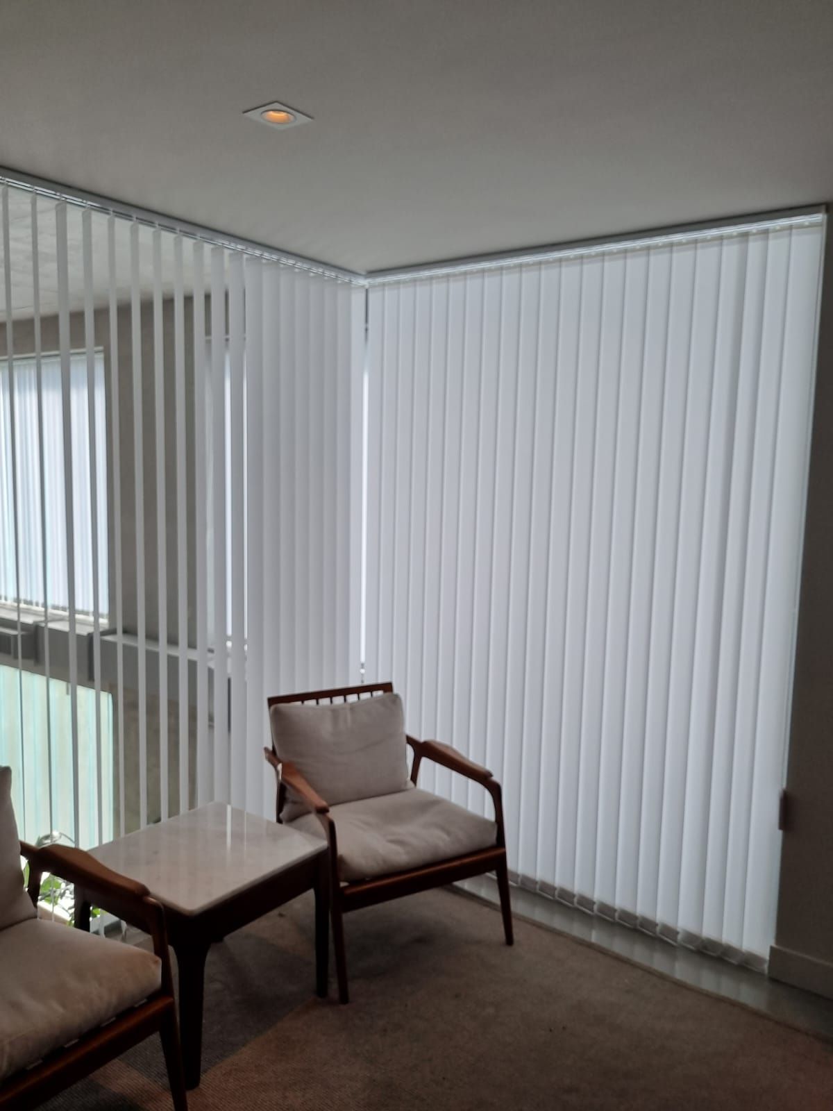Habitación vacía con persianas verticales, dos sillas de madera y una mesita junto a la ventana.