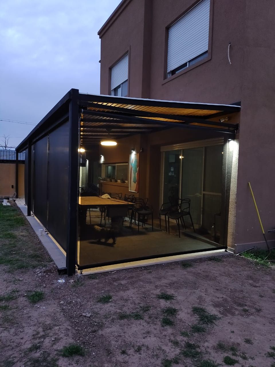 Patio con paredes de cristal anexo a la casa al anochecer, con iluminación interior cálida y una mesa en el interior.