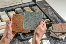 Hands holding two square terrazzo tile samples over a display of assorted tiles