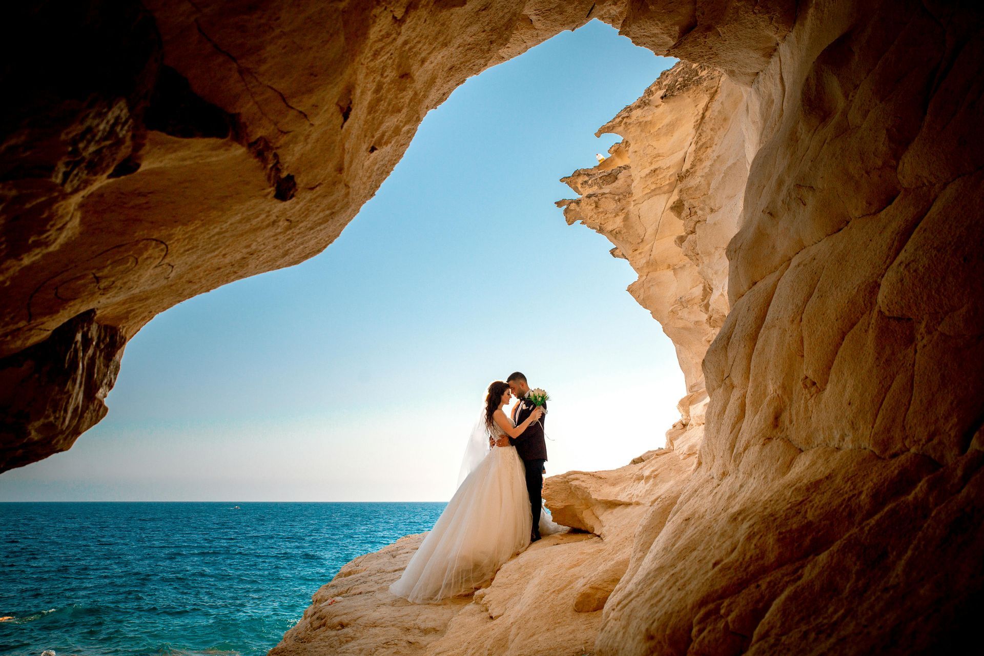 A bride and groom are kissing in a cave overlooking the ocean.