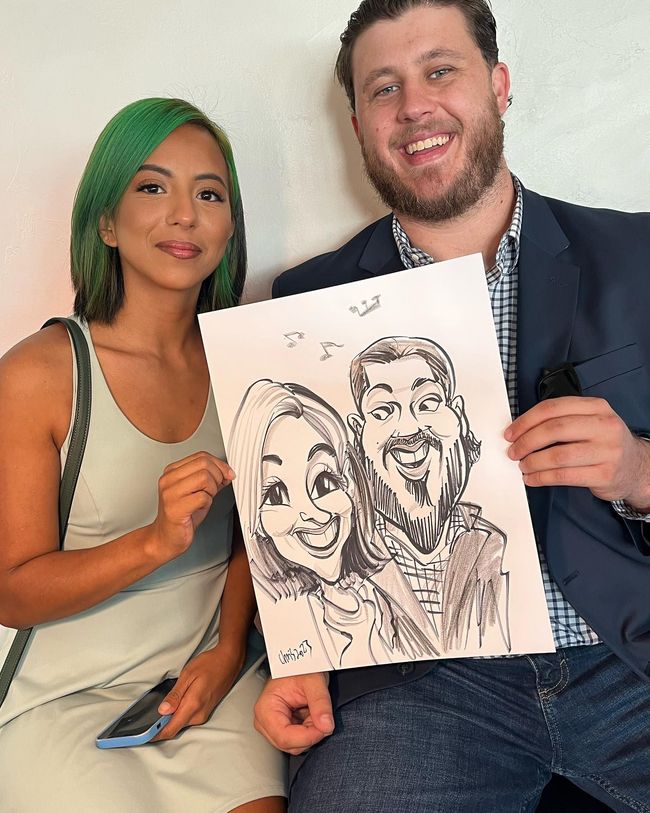 Two people holding a caricature drawing of themselves, smiling on a couch.