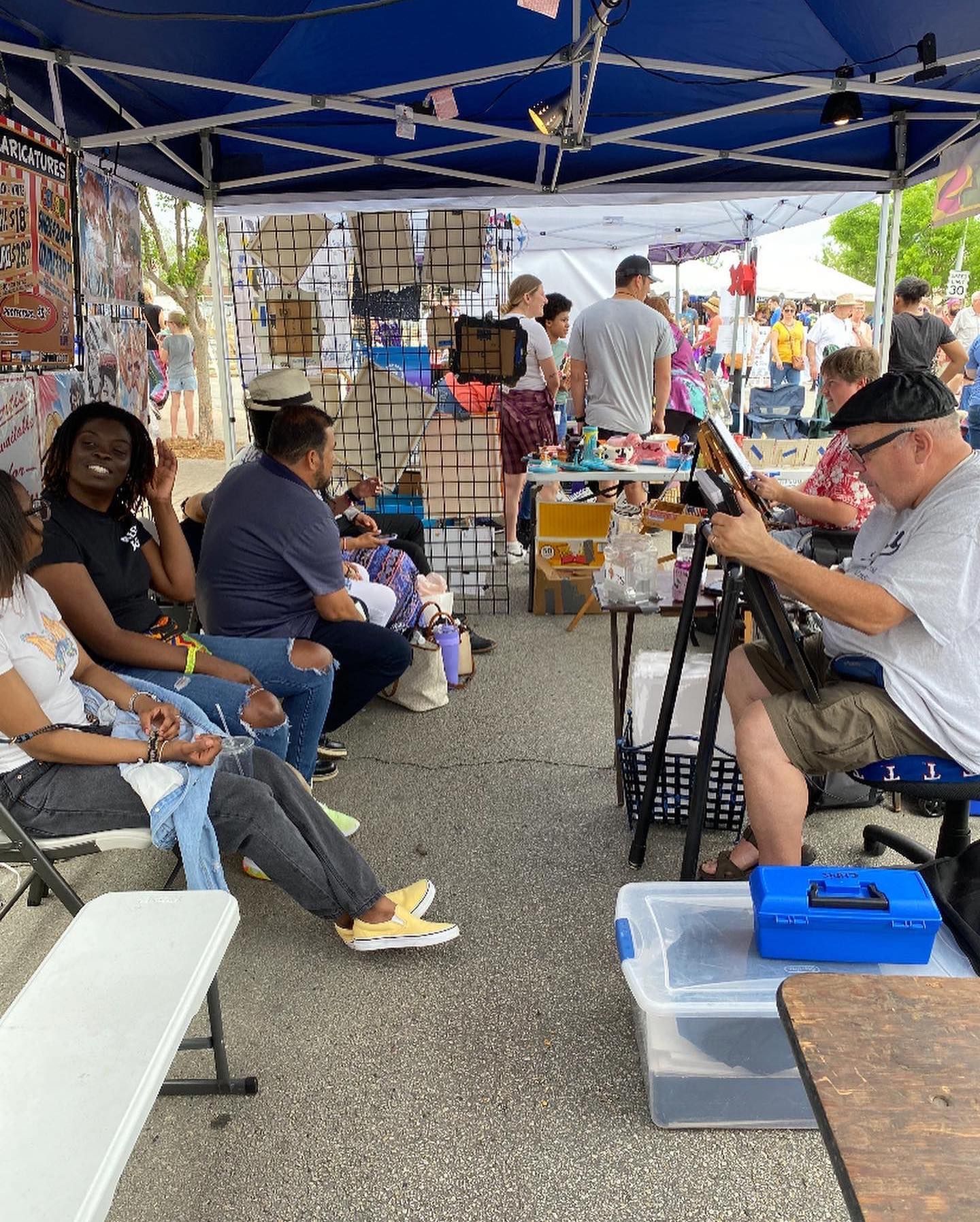 People seated under a blue canopy at an outdoor market stall, browsing items on display tables.