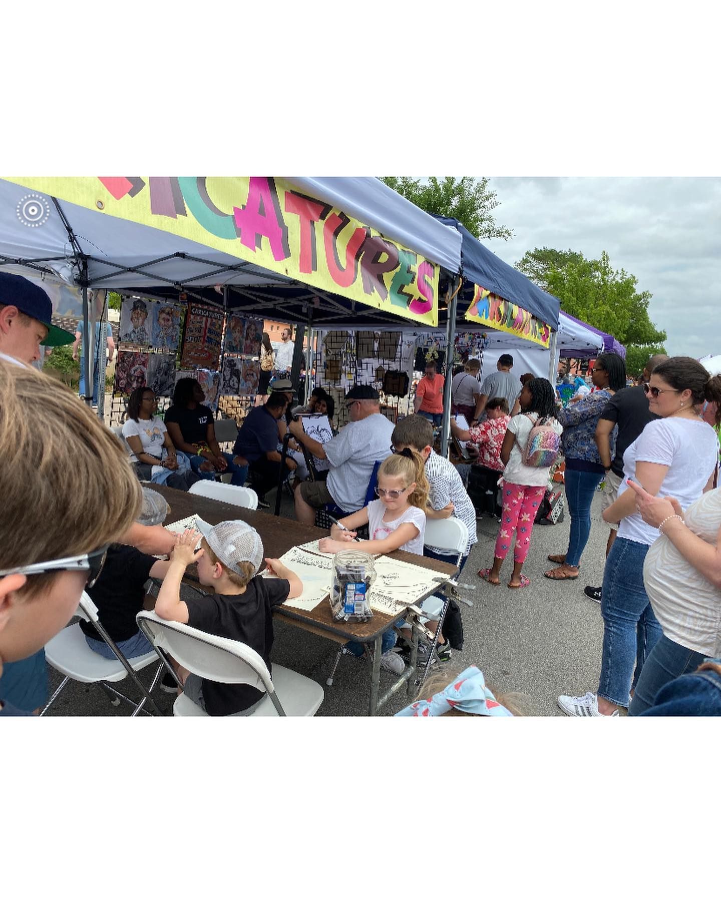 Crowded outdoor food stall under a colorful canopy, with people lined up and sitting at nearby tables.