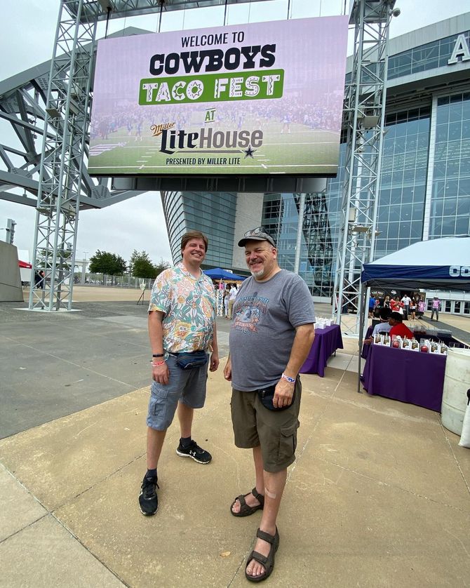 Two men posing under a Cowtown Taco Fest sign outside a glass building.