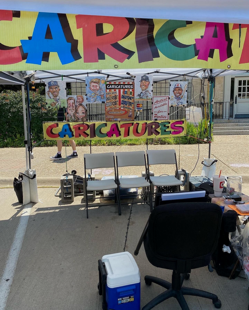 Outdoor caricature booth with colorful “CARICATURES” signs, chairs, and drawing supplies under a tent
