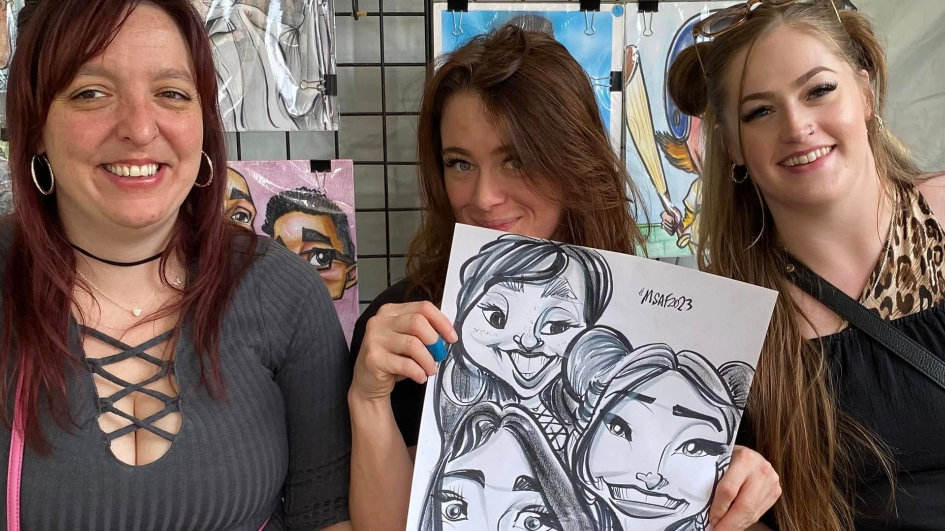 Two adults and a child holding colorful caricature portraits at an outdoor fair booth