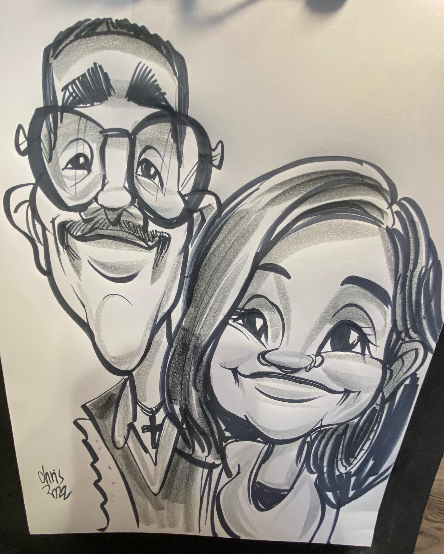 Black-and-white caricature drawing of two smiling people, one with glasses and a mustache, on paper