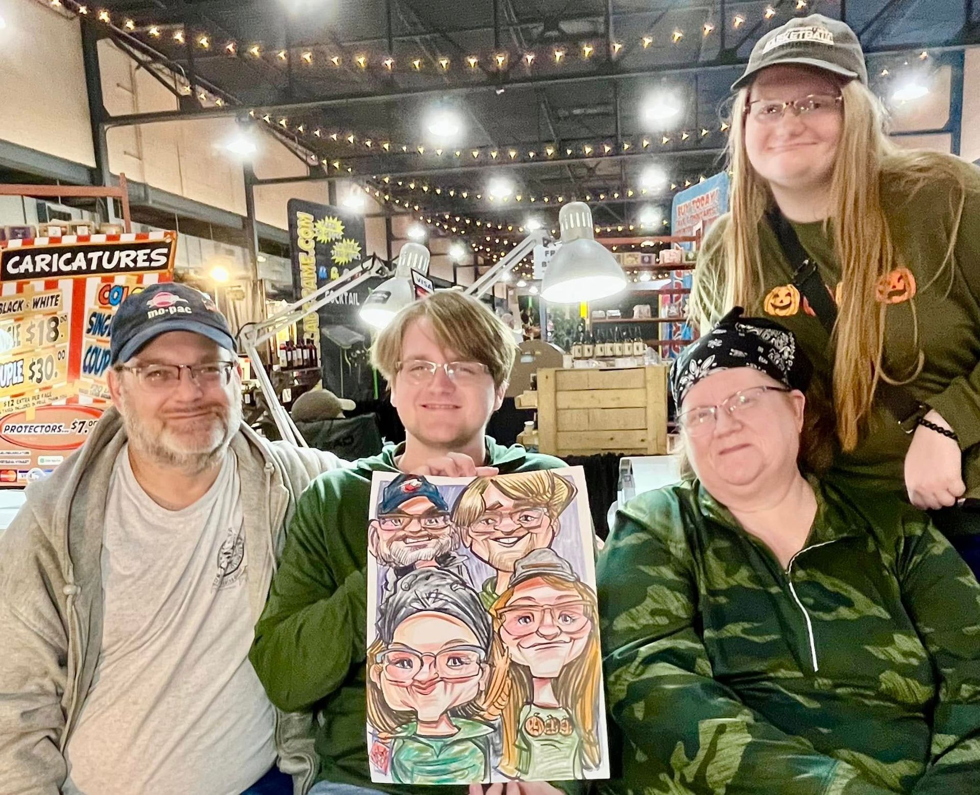Four people pose at a carnival booth, one holding a colorful caricature portrait, under string lights.