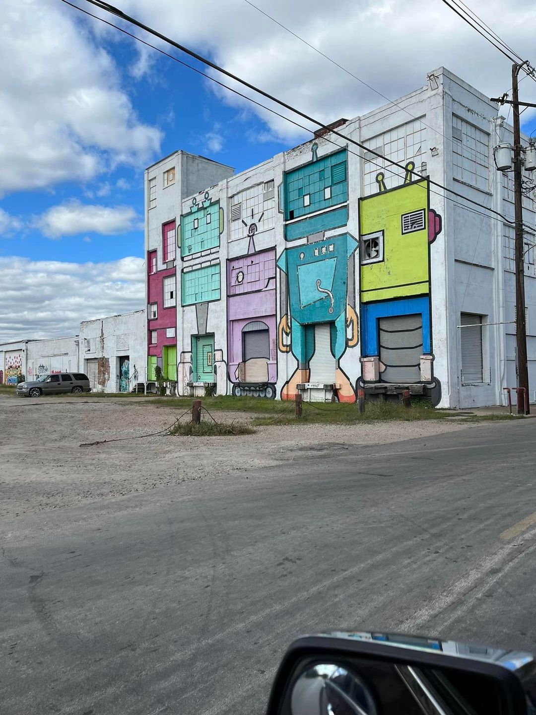 Colorful mural on a gray building beside a dirt road under a cloudy sky, with car hood in foreground