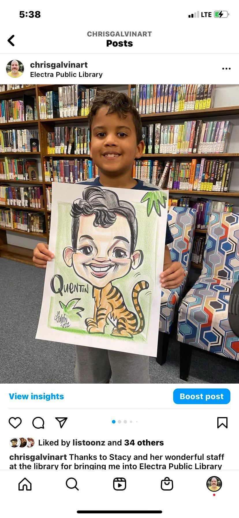 Child holding a caricature drawing in a library, smiling in front of shelves of books.