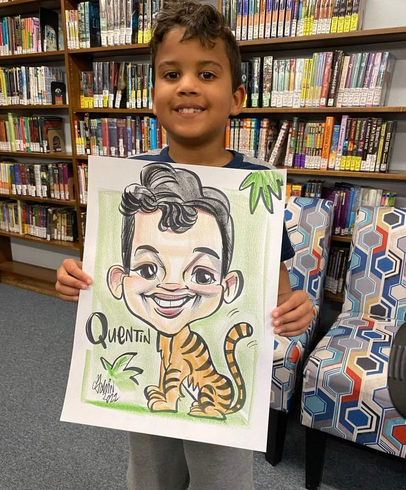 Child holding a colorful caricature drawing in a library, with bookshelves in the background.