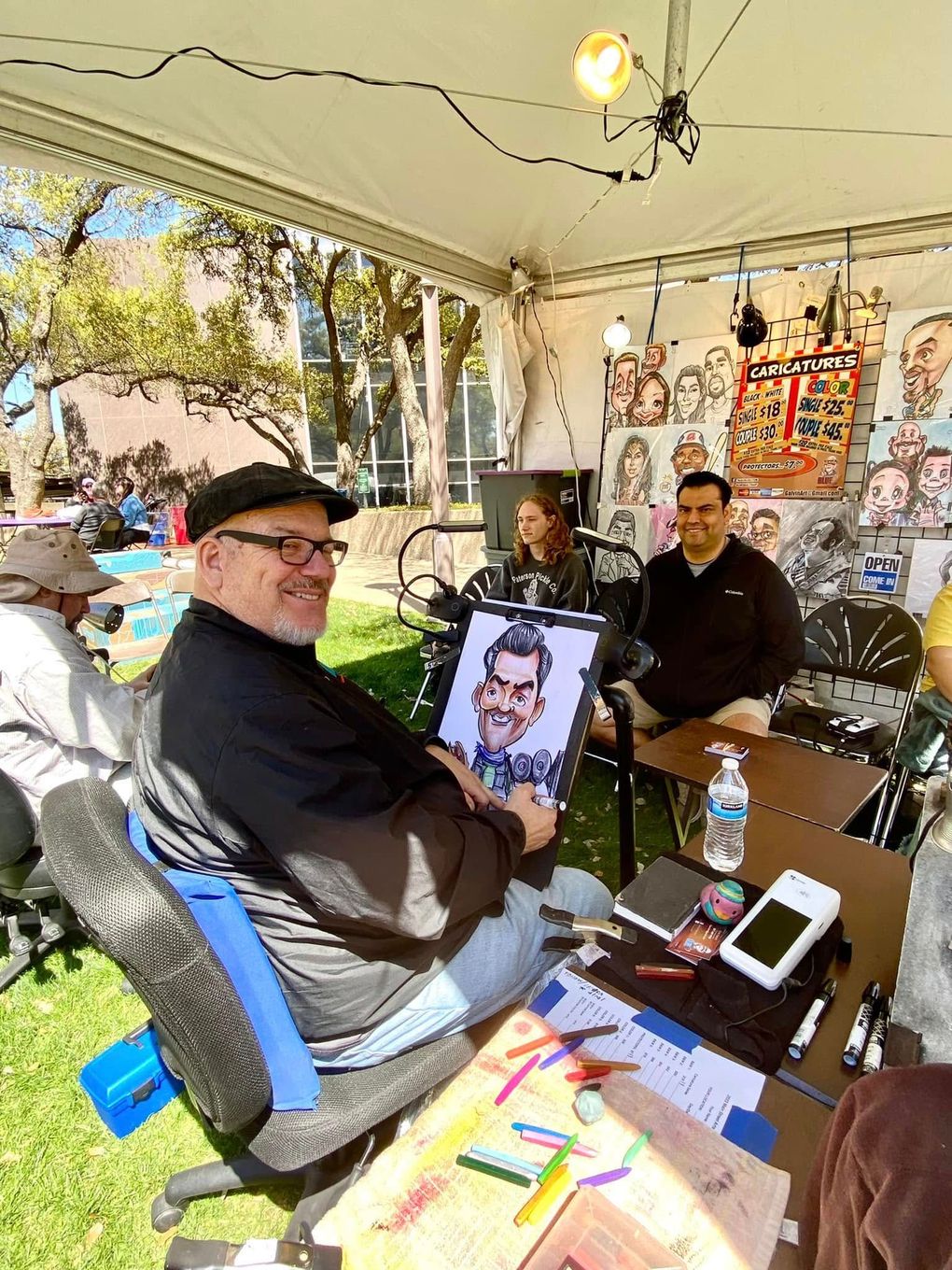 People seated under a white tent at an outdoor art booth, with colorful drawings displayed on the table.