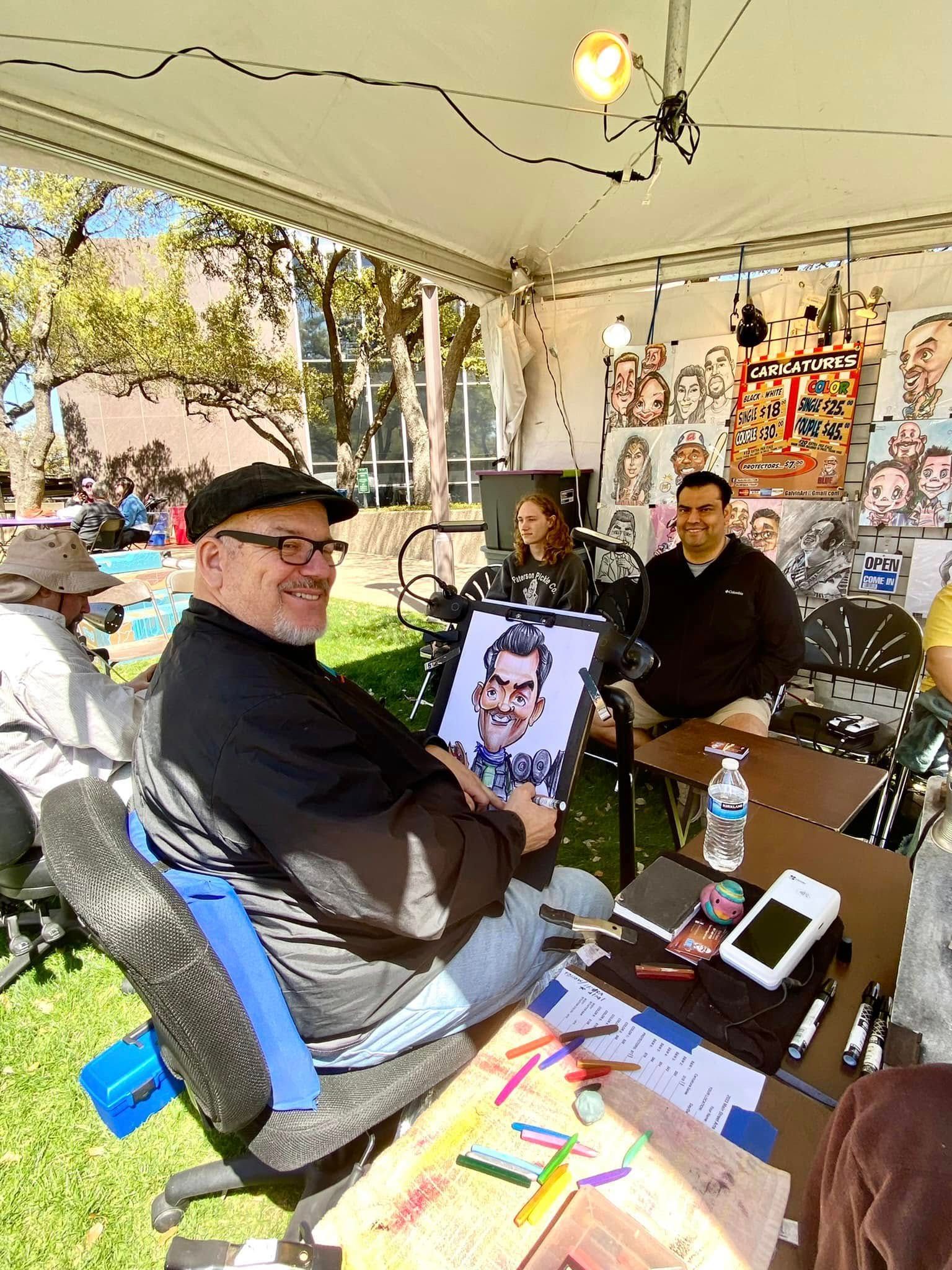 People sitting under a white tent at an outdoor art booth, with sketches and supplies on a table.