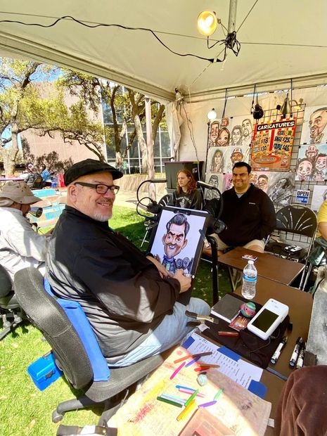 Man sitting under a white tent at a table with colorful art supplies and a portrait sketch nearby