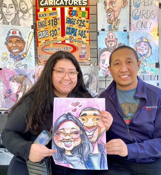 Two smiling people hold a caricature portrait in front of a wall of colorful caricature drawings.