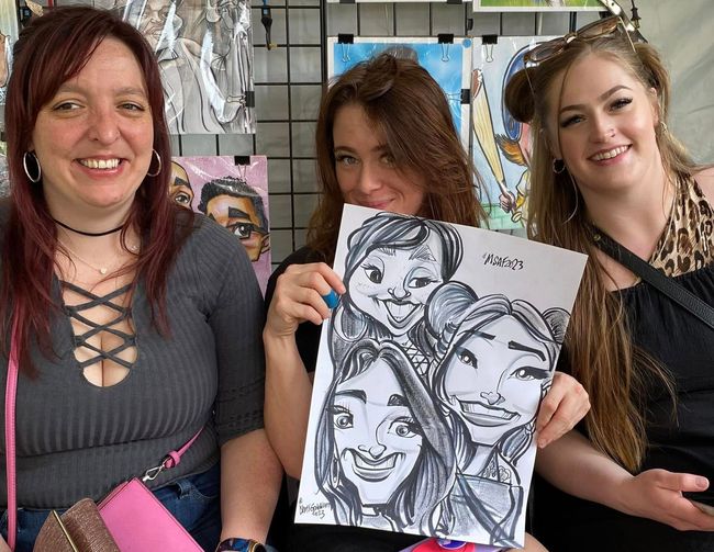Three smiling people at an outdoor market, one holding a black-and-white caricature drawing.