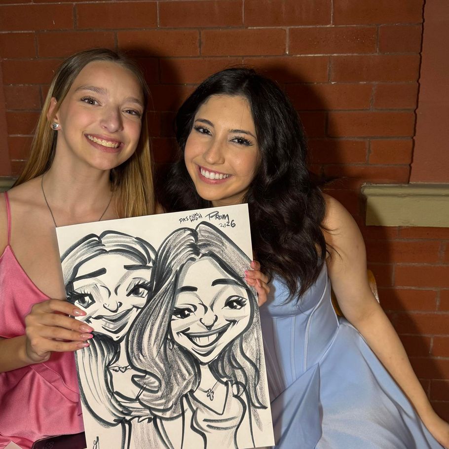 Two smiling friends holding a caricature drawing, standing before a brick wall.