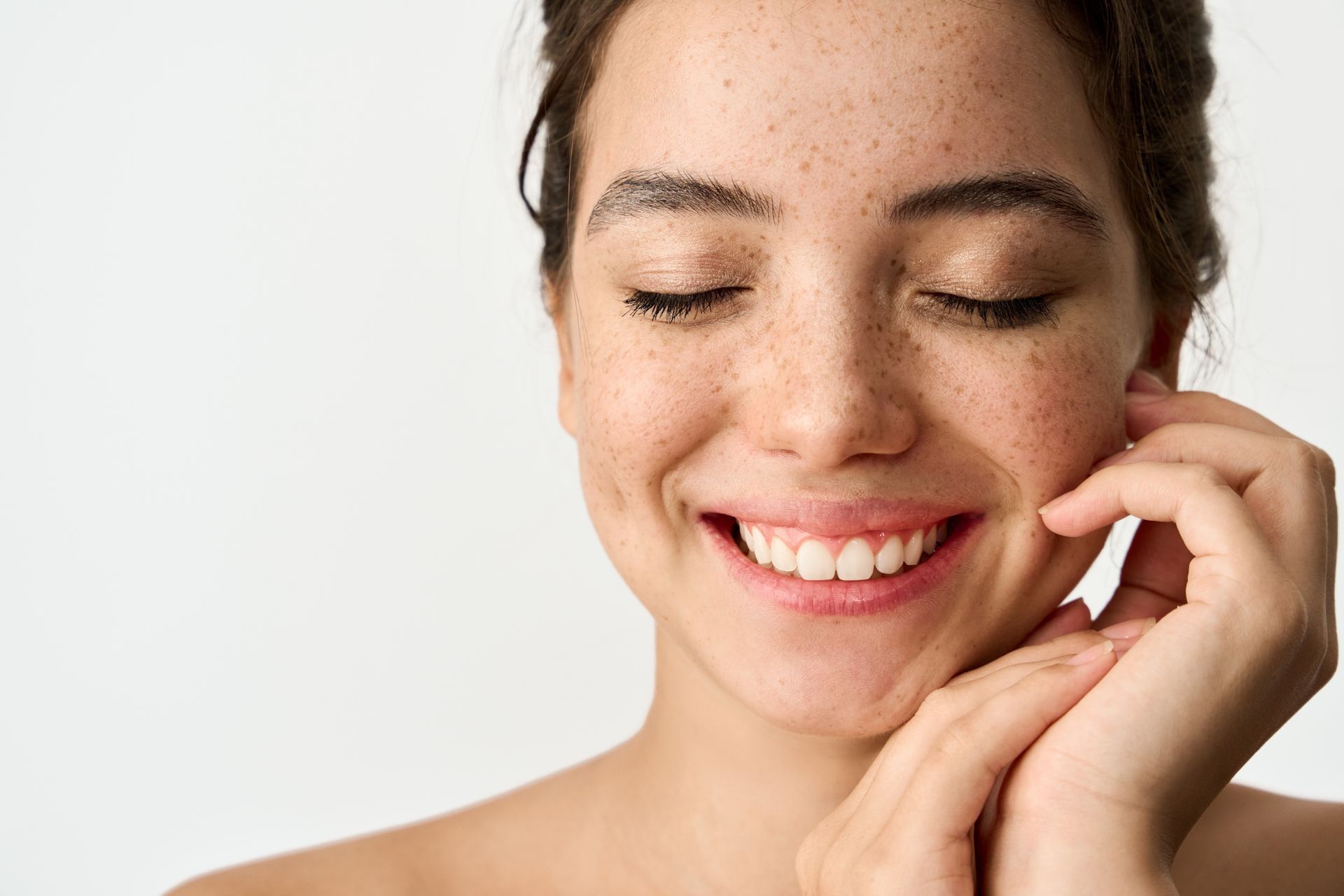 A woman with freckles is smiling with her eyes closed and touching her face.