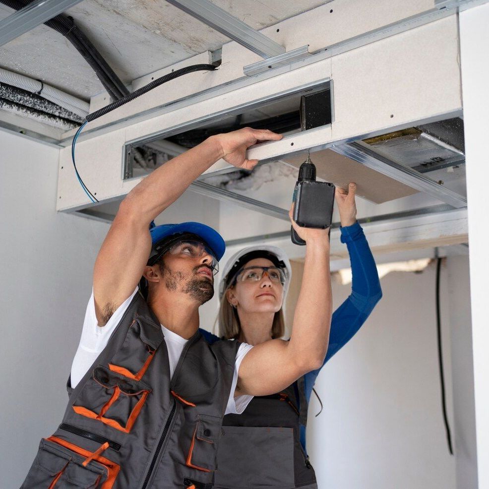 Two people installing a drywall ceiling with a drill; one wearing a blue hat, the other a hard hat.
