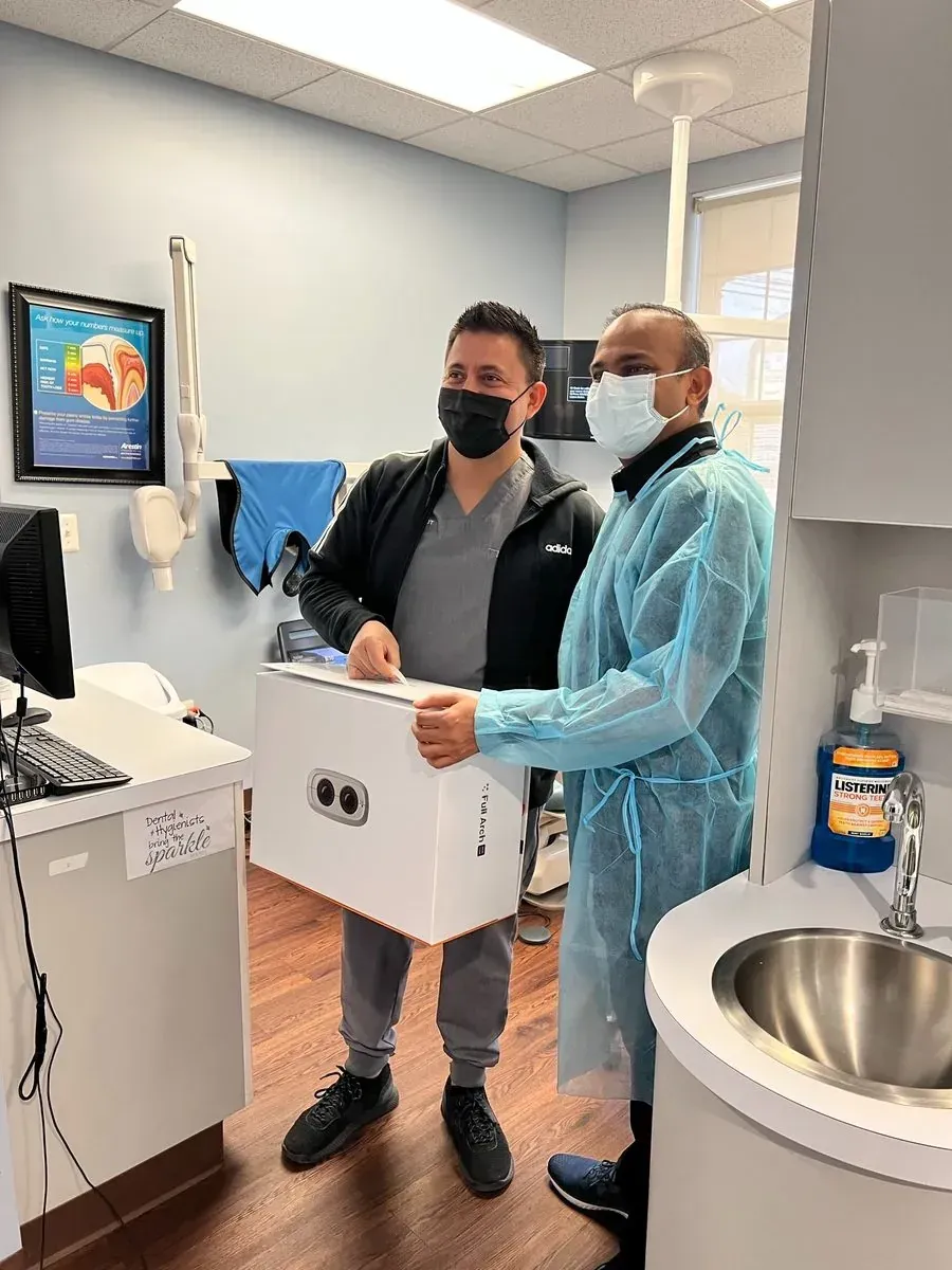 Two people in masks and gowns in a clinic, holding a white cooler by a sink.