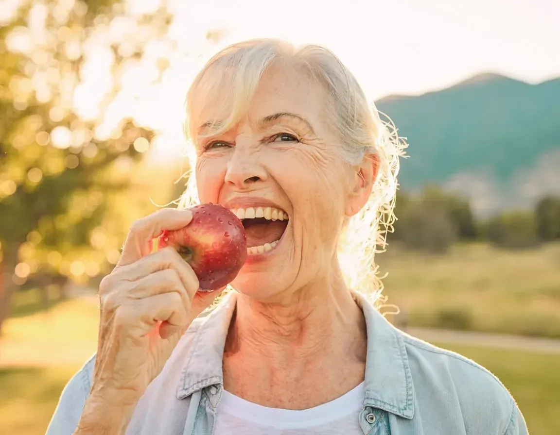 Woman holding a red apple and smiling outdoors in warm sunlight, with mountains in the background