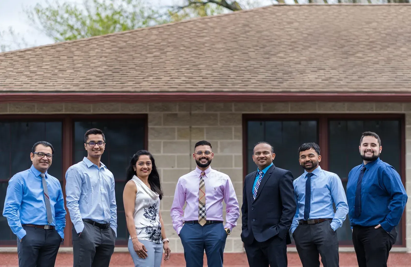 Seven people standing together outside a brick building, smiling in business-casual attire.