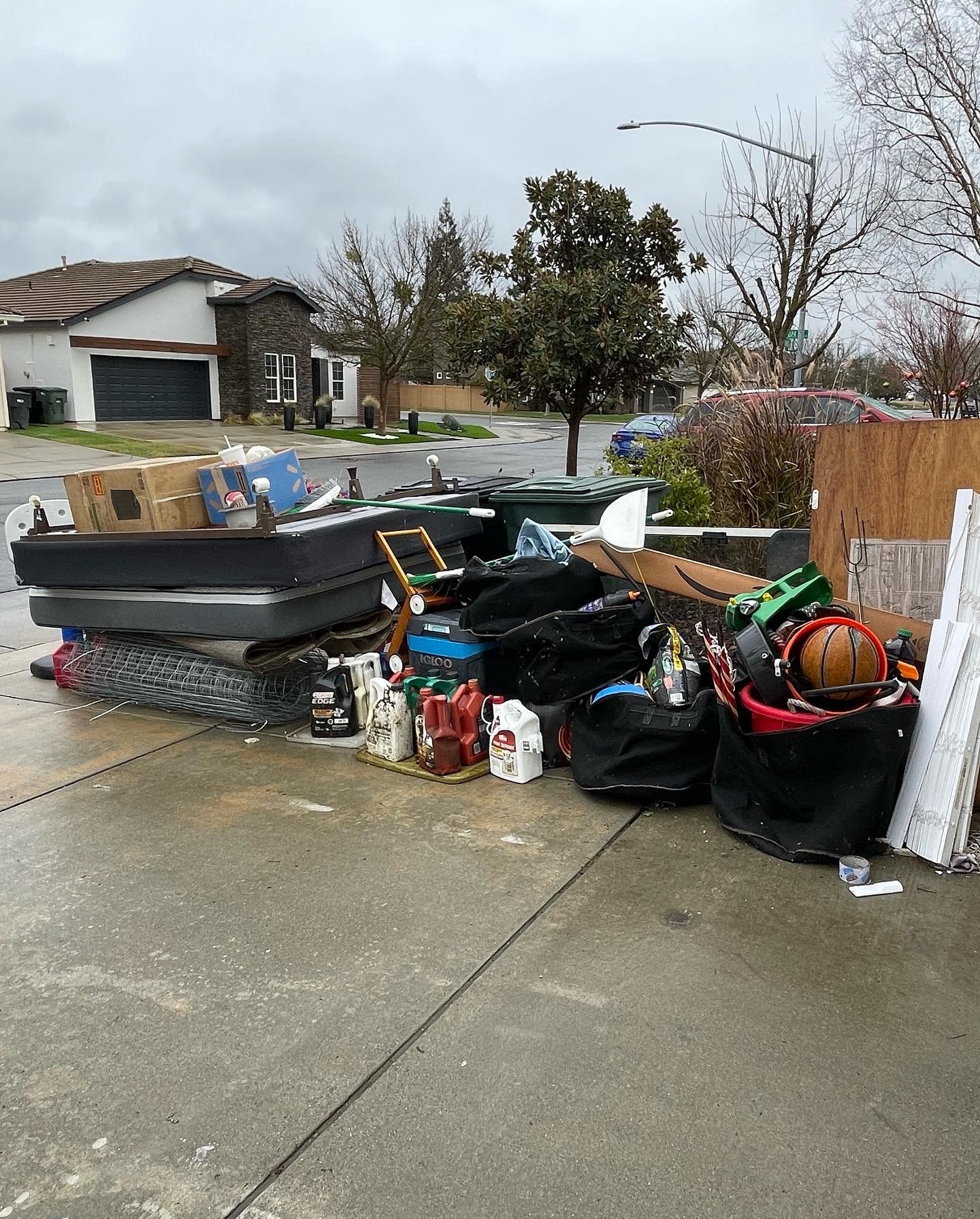 A large, messy pile of household trash, including mattresses, boxes, and bags, sits on a curb in a residential neighborhood.
